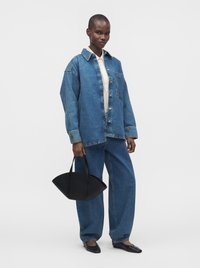 Femme portant une veste en jean bleue oversize et un jean large assorti, tenant un sac à main structuré noir, debout sur un fond blanc.