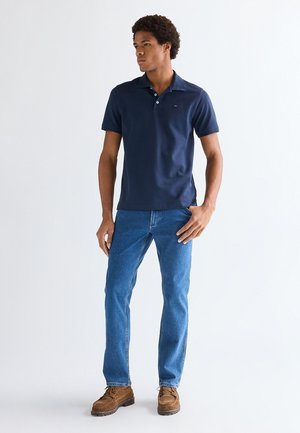 Νέος άνδρας στέκεται, φοράει μπλε σκούρο κοντομάνικο πόλο πουκάμισο, μπλε τζιν και καφέ κορδονένια παπούτσια σε λευκό απλό φόντο.