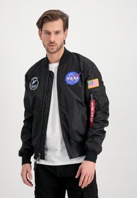 Black bomber jacket with NASA logo, American flag patch, zipper pockets, ribbed cuffs, and a red tag reading "REMOVE BEFORE FLIGHT."