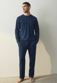 Marineblauw langemouwen shirt met een borstzak, gecombineerd met bijpassende elastyke taillebroek, beiden gemaakt van zachte stof; blote voeten zichtbaar.