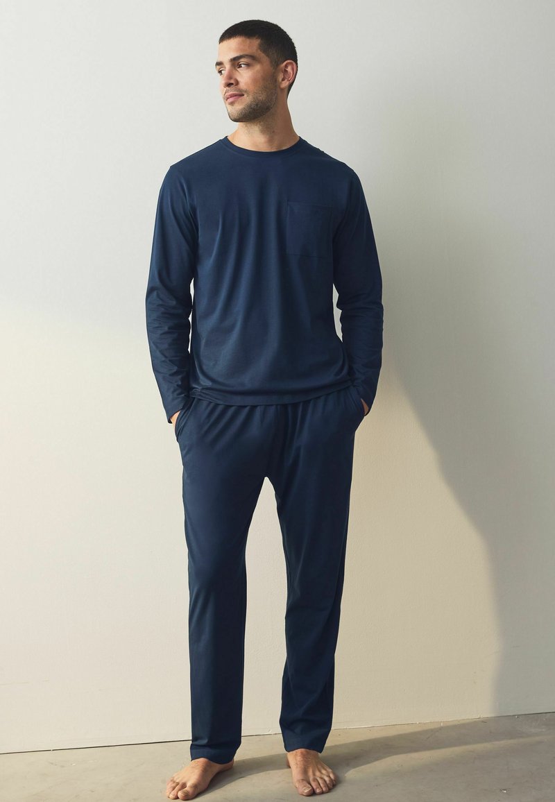 Marineblauw langemouwen shirt met een borstzak, gecombineerd met bijpassende elastyke taillebroek, beiden gemaakt van zachte stof; blote voeten zichtbaar.