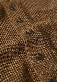 Cardigan marron en tricot avec une texture côtelée, comportant trois boutons sombres sur le devant. Le tissu brille légèrement sous la lumière.