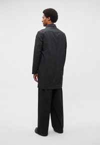 Fekete, egyenes szabású hosszú kabát, gallérral, gombos záródással és nagy zsebekkel. Lazas fekete nadrág fölött viselve.