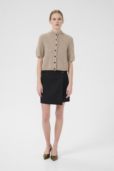 Cardigan en tricot beige à manches courtes avec un col haut et des boutons noirs, associé à une jupe portefeuille noire et des escarpins verts à bout pointu.