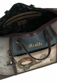 Multikleurige handtas met een gestructureerd oppervlak, voorzien van een bruin en blauwe design, gouden logo en een decoratieve charm. Met rits aan de bovenkant.