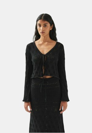 Donna con lunghi capelli scuri che indossa un cardigan nero in maglia con lacci frontali e una gonna in maglia abbinata a vita alta su uno sfondo semplice.