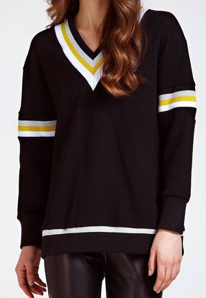 Žena v černém svetr s výstřihem do V a bílým a žlutým proužkováním na límci, rukávech a lemu, v kombinaci s černými kalhotami.