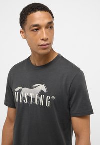 Camiseta de algodón gris oscuro con un gráfico de un caballo blanco y el logo "MUSTANG". Cuello redondo, corte regular y mangas cortas.