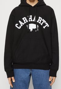 Černá mikina s kapucí s bílým nápisem „CARHARTT“ a grafikou bizona. Má přední kapsu a nastavitelné šňůrky na kapuci.