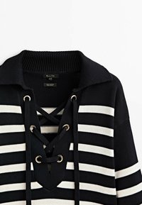 Pull en maille à rayures noires et blanches avec un grand col et un col en V lacé avec des cordons noirs passant à travers des œillets argentés.