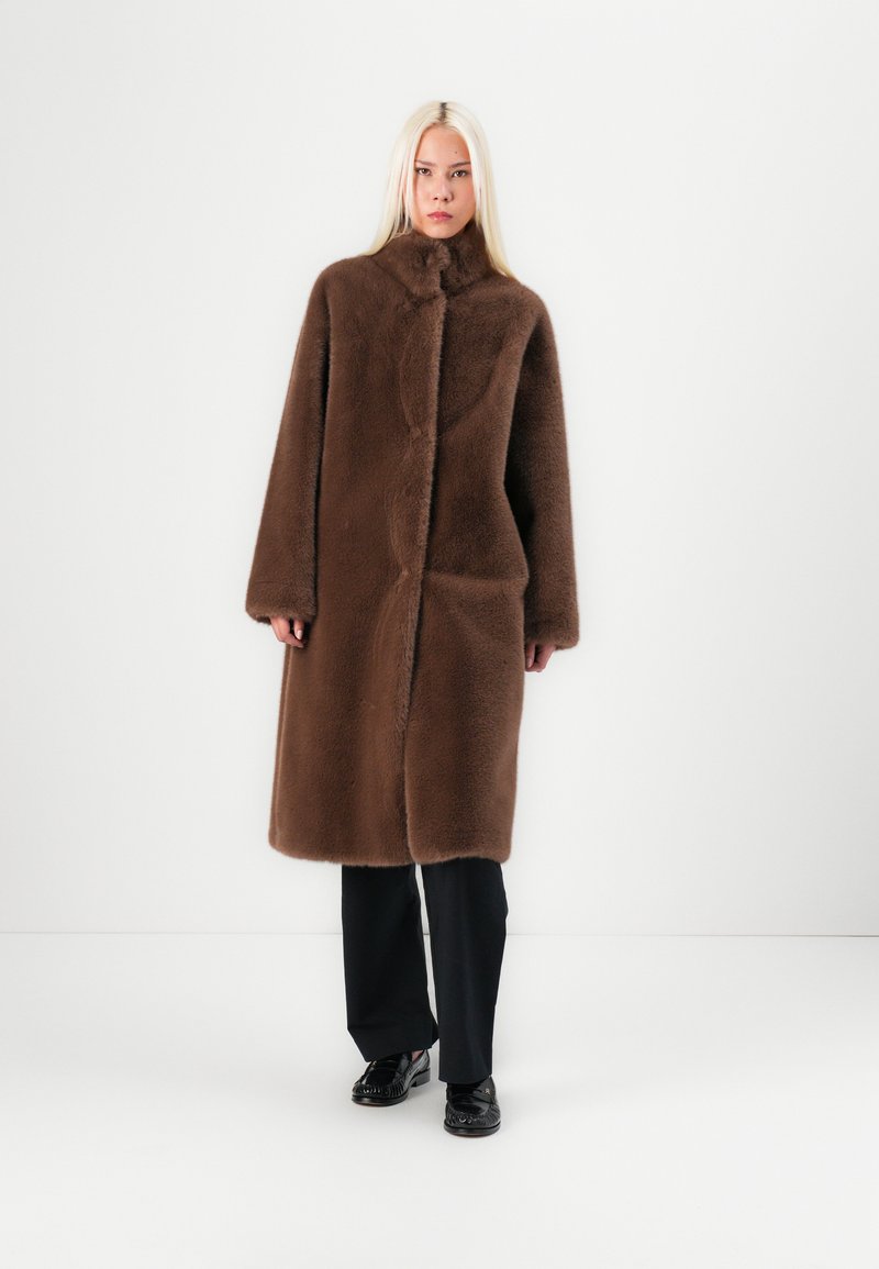 Manteau en fausse fourrure marron avec un col montant, des manches longues et une coupe droite. Dispose de poches latérales et d'une texture veloutée. Pantalons et chaussures noirs.