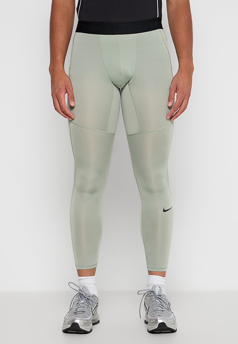 Hombre vistiendo mallas de compresión Nike Pro verde claro, camiseta negra, calcetines blancos y zapatillas Nike plateadas de pie contra un fondo blanco.