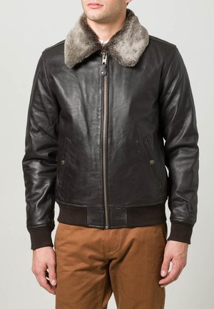 Man in een donkerbruin leren jack met een grijze bontkraag en een bruine broek, staand tegen een vlakke lichte achtergrond.
