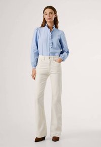 Svetlo modra srajca z gumbi in volančastim izrezom, kombinirana z belimi razširjenimi kavbojkami in rjavimi čevlji z visokimi petami, ki ustvarjajo oprijeto silhueto.