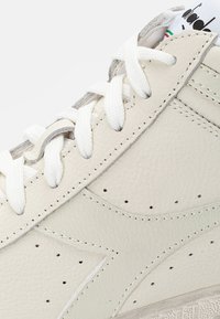 White leather sneaker featuring a textured finish, flat white laces, perforations, and a subtle gray accent near the top.