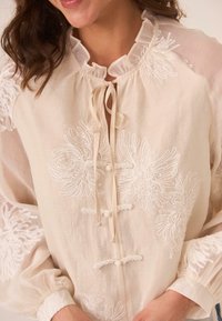 Blusa color crema con cuello con volantes, diseño floral bordado en los hombros, cierre con lazo y mangas amplias con acentos de botones.