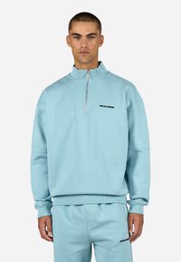 Gaiši zils zip-up džemperis ar ribota tipa piedurknēm un apakšmalu, kuram uz krūtīm ir melns "PEGADOR" logo un augsta apkakles dizains.