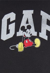 Camiseta negra con un gran logo plateado "GAP" y un ratón de dibujos animados con pantalones cortos rojos y zapatos amarillos, exhibido en una pose juguetona.
