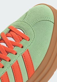 Zelene suède tene stilizirane s narančastim vezicama i prugama, s teksturiranim dizajnom i kontrastnom smeđom gumiranom petom.