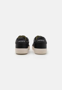 Zapatillas de cuero negro con suela de goma crema. Incluyen un cuello y lengüeta acolchados, con la marca "LACOSTE" en el talón.