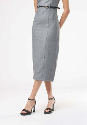 Donna che indossa un vestito midi grigio aderente con una sottile cintura nera e sandali neri con tacco alto su sfondo bianco.