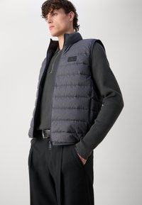 Gepolsterte Weste in dunkelblau, mit hohem Kragen, strukturiertem Stoff und einem Logopatch auf der Brust. Trägt man über einem schwarzen Langarmoberteil.