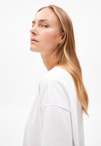 Weißes, übergroßes, langärmliges Baumwollhemd mit Rundhalsausschnitt. Glatte Textur und entspannter Schnitt. Das Model hat glattes Haar und ist leicht zur Seite gewandt.
