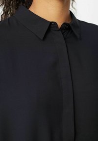 Schwarzes Hemd mit Kragen aus glattem Stoff. Verfügt über eine Knopfleiste und einen sauberen Abschluss um den Kragen und die Schultern.