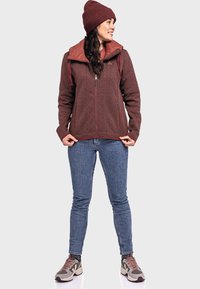 Rotbraune Jacke mit strukturierter Oberfläche, grauen Akzenten, Reißverschlusstaschen, kombiniert mit Jeans und gemusterten Sneakers, abgerundet mit einem passenden Beanie.