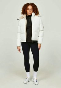 Vit puffjacka med dragkedja fram och beige detaljer, bärs över en svart topp, ihop med svarta leggings och vita sneakers.