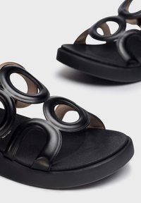 Sandalias negras hechas de material texturizado, con recortes circulares, una suela plana y una plantilla acolchada para mayor comodidad.