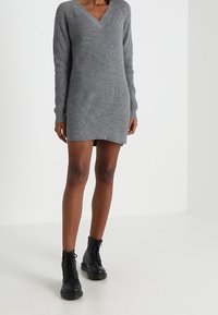 Robe grise en tricot côtelé à col en V, avec manches longues et accents diagonaux à l'ourlet, portée avec des bottes noires à lacets.