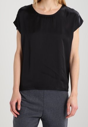 Persona que lleva una blusa negra lisa de manga corta y pantalones grises, de pie con los brazos relajados a los lados frente a un fondo gris.