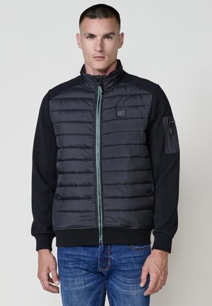 Pletená čierna bunda s polstrovanými rukávmi, predným zipsom a malým logom. Disponuje kontrastným zeleným detailom na zips a bočnými vreckami.