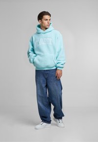 Light blue fleece hoodie with white “KANI” text, front pocket, and hood, paired with loose blue jeans and white sneakers.
