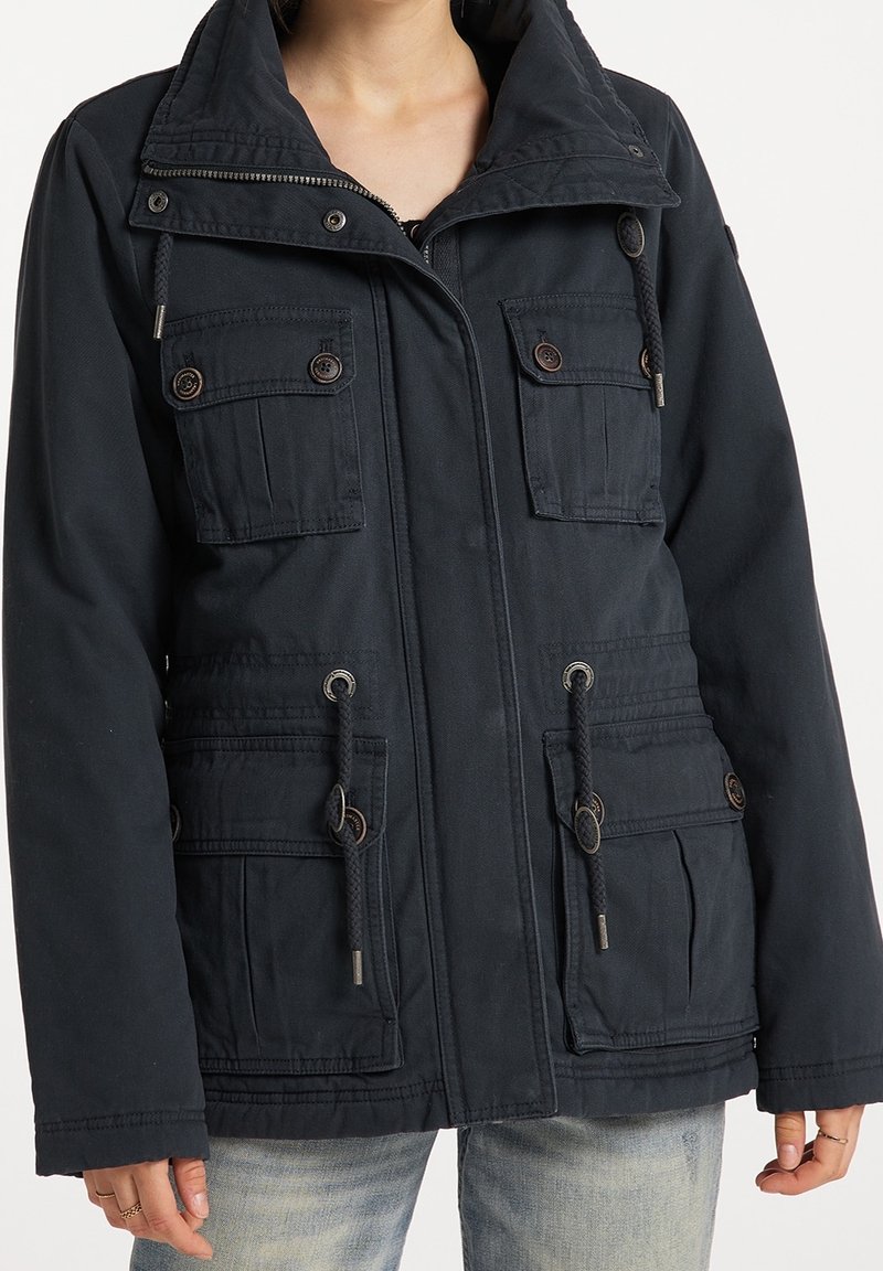 Personne portant une veste à capuche noire avec fermeture éclair frontale, poches à rabat boutonnées et taille à cordon de serrage, associée à un jean bleu clair.