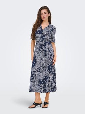 Femme aux longs cheveux bruns portant une robe midi boutonnée bleu marine et blanc à imprimé cachemire avec des manches courtes et des sandales à talons noires.