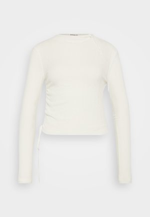 Long-sleeve, white ribbed top with a crew neckline and adjustable side ruching. Features a tie detail on the left shoulder.