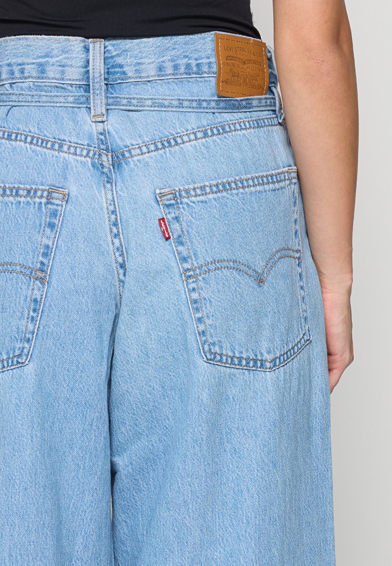 Jean en denim bleu clair à taille haute, avec deux poches arrière et un patch en cuir marron au niveau de la taille.