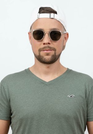 Uomo che indossa una maglietta verde a scollo a V, occhiali da sole rotondi e un cappellino da baseball bianco portato al contrario, guarda dritto davanti a sé.
