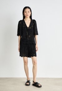 Abito in pizzo nero con scollo a V, maniche a palloncino corte e orlo ondulato, caratterizzato da intricati motivi a taglio circolare e un fiocco sul davanti.
