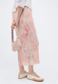 Γυναίκα που φοράει φούστα με ροζ και μπεζ λουλουδένιο πιέτες, λευκό πουκάμισο με ρυτιδωμένα μανίκια, μπεζ και λευκά αθλητικά παπούτσια, κρατώντας μια μικρή τσάντα χειρός σε γυμνό ροζ χρώμα.