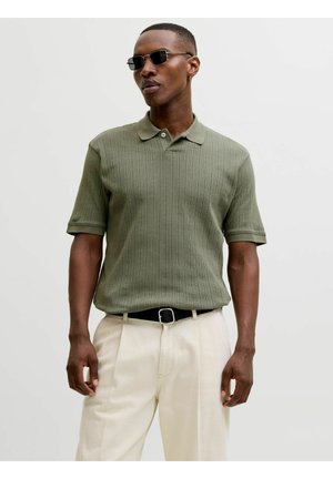 Muž so stmavými slnečnými okuliarmi, olivovo zeleným krátkym pleteným polokošeľom, béžovými nohavicami a čiernym opaskom, stojí pred jednofarebným svetlým pozadím.