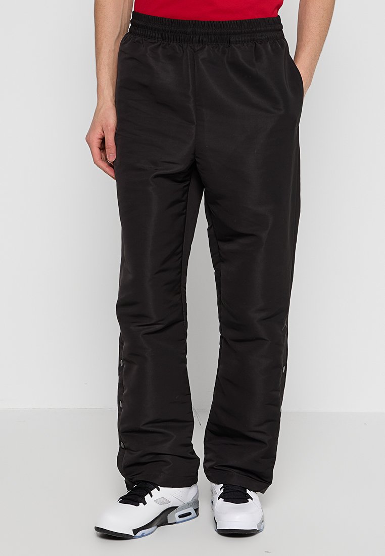 Schwarze gepolsterte Hosen mit elastischem Bund, seitlichen Taschen und Druckknopfverschlüssen an den Knöcheln, kombiniert mit weißen Sportschuhen.