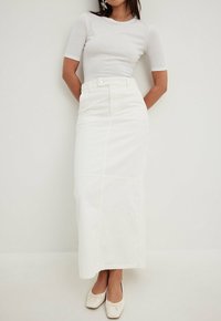 Jupe maxi en coton blanc avec taille cintrée, dotée d'une fermeture à boutons sur le devant et de coutures verticales, associée à un haut ajusté gris clair et des chaussures crème.