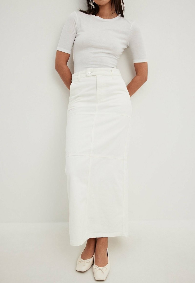 Jupe maxi en coton blanc avec taille cintrée, dotée d'une fermeture à boutons sur le devant et de coutures verticales, associée à un haut ajusté gris clair et des chaussures crème.