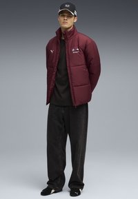 Veste matelassée bordeaux avec logos BMW et Puma, col montant, associée à un t-shirt noir, un jean noir à jambes larges et des chaussures noires.