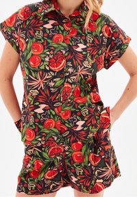 Combinaison à manches courtes en tissu foncé, avec un imprimé vif de fruits et de feuillages. Boutons sur le devant et poches sur le short.