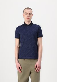 Mørkeblå polo skjorte med mønsterdesign, korte ermer og mørk krage. Modellen bruker den sammen med khakibukser, stående mot en hvit bakgrunn.