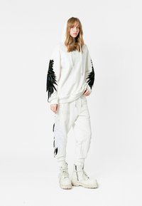 Sudadera blanca con diseños de plumas negras en las mangas, combinada con joggers blancos con acentos bordados y botas de combate blancas.